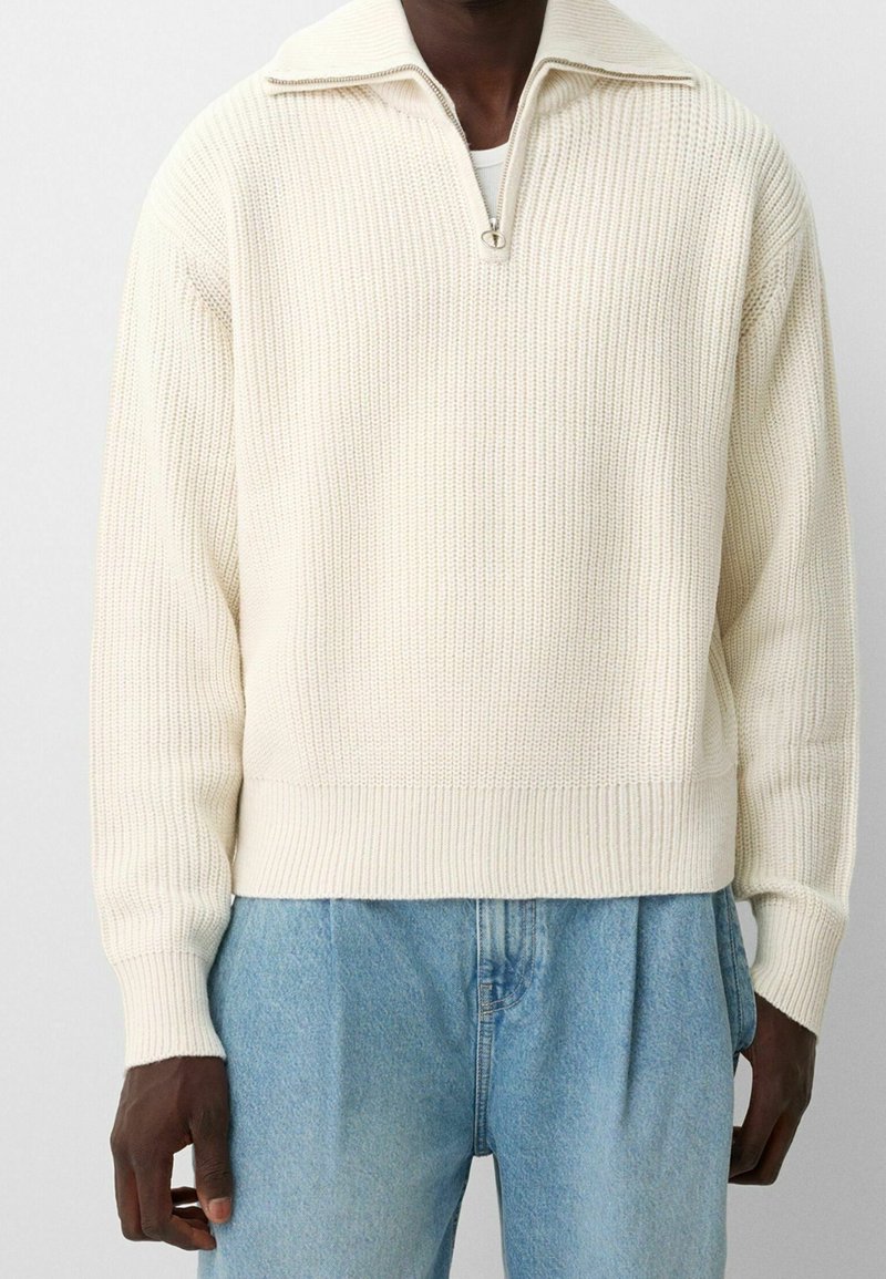 Homme portant un pull en maille côtelée couleur crème avec col à demi-fermeture éclair et jean taille haute bleu clair, debout devant un fond uni.