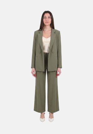Donna in piedi, indossa un tailleur pantalone verde oliva a gamba larga con blazer abbinato e una canotta in raso color crema, tacchi bianchi a punta.