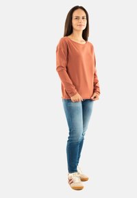 Donna con lunghi capelli scuri che indossa un maglione color ruggine, jeans blu e scarpe da ginnastica bianche, in piedi contro uno sfondo chiaro e uniforme.