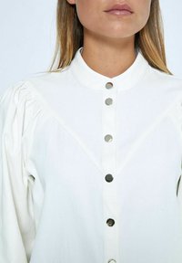 White long-sleeve shirt with a high collar, featuring silver snap buttons down the front and textured puff sleeves. Smooth cotton fabric.