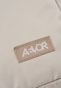 Beige Stoffoberfläche mit einer strukturierten Webart, die ein gesticktes rechteckiges Schild mit der Aufschrift "A=VOR" in weißer Schrift aufweist.