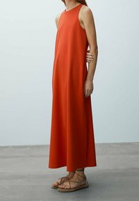 Robe maxi sans manches rouge à coupe ample, avec un col haut et des fentes latérales. Portée avec des sandales à lanières beiges, sur un fond neutre.