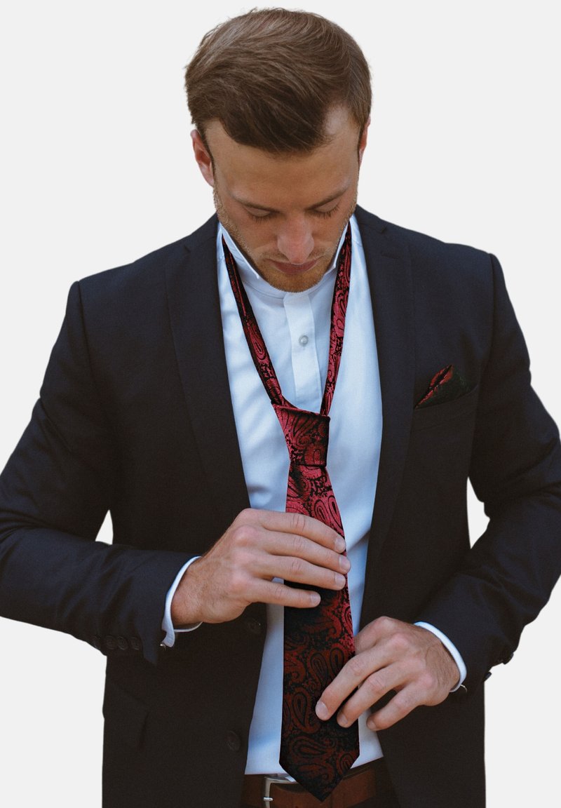 Uomo in abito scuro e camicia bianca che guarda verso il basso mentre sistema una cravatta rossa e nera con fantasia.