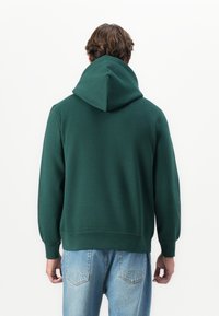 Man with wavy brown hair wearing dark green hooded sweatshirt and light blue jeans, standing facing away against plain white background.
