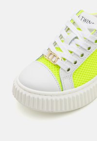 Sneaker blanche avec panneau en maille jaune fluo, lacets blancs, accent matériel doré avec logo, et semelle en caoutchouc blanche texturée.