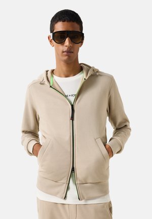 Mand iført store mørke solbriller og en beige lynlåshætte trøje med hænderne i lommen mod en ensfarvet baggrund.