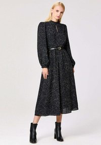 Robe noire avec de longues manches bouffantes, un détail en trou de serrure, un motif parsemé d'or, une taille ceinturée et une jupe fluide. Associée à des bottines noires.
