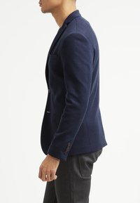 Mörkblå blazer i texturerad tyg, med en enda ficka, notch-lapel och detaljer med tre knappar på ärmarna. Skräddarsydd passform.