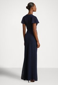 Μακρύ φόρεμα σε ναυτικό μπλε με φουσκωτά μανίκια, εφαρμοστή μέση και διάφανο περίβλημα. Κλείνει στο πίσω μέρος με κρυφό φερμουάρ. Λείο υφή.