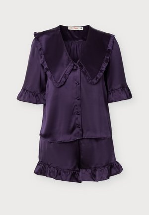 Mujer rubia de pie descalza, vistiendo una blusa de satén de manga corta y botones en color púrpura oscuro con cuello con volantes y pantalones cortos a juego con volantes en el bajo.