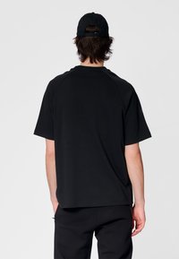 Svart t-shirt i bomull med korta ärmar, avslappnad passform, rund halsringning och raglanärmar, visad bakifrån mot en enfärgad bakgrund.