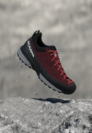 Scarpa da trekking bordeaux e nera con lacci rossi, fotografata in aria sopra un terreno roccioso con particelle di polvere sottostanti.