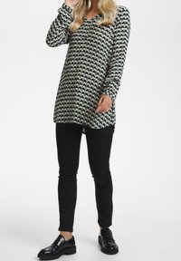 Tunique longue à motifs verts, bleu marine et blancs en formes géométriques ; associée à un pantalon noir ajusté et des mocassins noirs. Design à encolure en V.