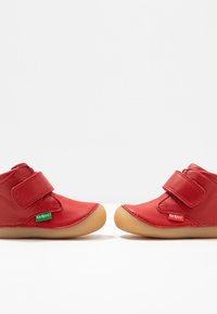 Chaussures pour enfants en cuir rouge avec brides à scratch et semelles en caoutchouc beige ; étiquettes de logo sur le côté. Texture lisse, forme arrondie.