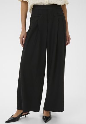 Donna che indossa pantaloni neri a vita alta, gamba larga, con tasche laterali e scarpe nere con tacco kitten appuntito.