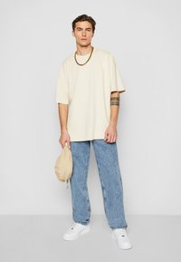 Lys beige oversized t-skjorte, avslappet passform. Blå denimjeans, lett avsmalnende. Hvite joggesko. Holder en beige glidelåspose. Enkel kjedehalskjede.