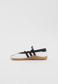 Sapatilha atlética de perfil baixo com um upper sintético branco, detalhes em preto, três tiras laterais e uma sola de borracha de cor caramelo. Apresenta um fecho de atacadores.