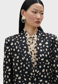 Crni sako s bež apstraktnim mrljama, uparen s bež košuljom koja ima crne točke. Zlatne i crne okrugle naušnice zaokružuju izgled.