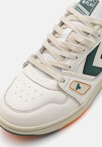 White leather sneaker with green accents, perforated toe, and beige laces. Features a patterned side patch and orange detailing on the sole.