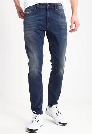 Vaqueros de denim en azul oscuro con un acabado ligeramente desvaído, diseño de pierna ajustada y detalles de costura en contraste. Combinados con zapatillas deportivas blancas.