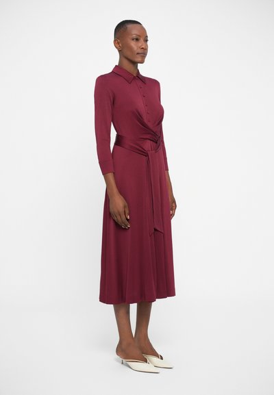 Burgundska haljina na dugih rukava s gumbima, s dizajnom omota i mašnom u struku, uparena s bijelim cipelama na rastezanje. Glatka tekstura tkanine.