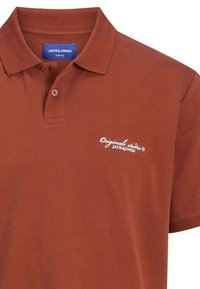 Polo marron en coton. Présente un col classique, des manches courtes et un logo blanc sur la poitrine. Deux boutons au niveau du cou.