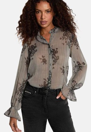 Femme portant un chemisier transparent à manches longues avec une broderie florale noire et des rayures verticales, assorti à un jean noir taille haute.