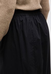 Άτομο που φοράει μπεζ πλεκτό πουλόβερ, σκουπισμένο μέσα σε μαύρη, ρυθμισμένη φούστα με ελαστική μέση, δείχνοντας την περιοχή της μέσης και των γοφών από πίσω.