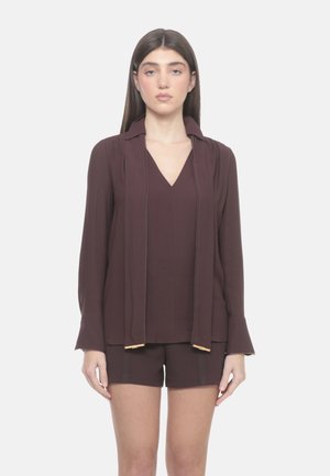 Giovane donna con capelli castani lisci che indossa una camicetta a maniche lunghe marrone scuro con lacci larghi al collo e pantaloncini abbinati, in piedi contro uno sfondo chiaro.