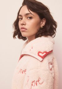 Puhast roza jakna z rdečim vezenjem "ljubezen" in srčastimi poudarki na ovratniku. Ima zadrgo in mehak, teksturiran material.