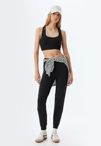 Schwarzes Sport-Crop-Top mit breiten Trägern, kombiniert mit eng zulaufenden schwarzen Hosen. Ein gebundenes gestreiftes Hemd sorgt für das gewisse Etwas, ergänzt durch weiße Sneaker.