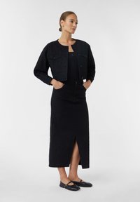 Black cropped denim jacket with button closure and two chest pockets, paired with a long black skirt featuring a front split. Black flat shoes.