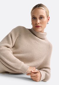 Gefütterter beiger Strickpullover mit hohem, geripptem Rollkragen, überschnittenen Schultern und strukturiertem Stoff. Lange, eng anliegende Ärmel runden das Gesamtbild ab.