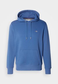 Modra kapucarica z žepi spredaj, vrvicami, rebrastimi manšetami in majhnim logotipom na levi strani prsi na enobarvnem ozadju.