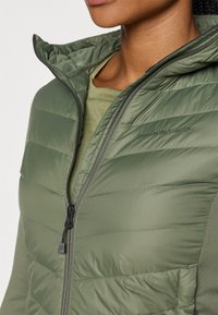 Giacca a vento imbottita verde con zip frontale, colletto alto e dettagli con logo. Presenta un'imbottitura orizzontale e una superficie liscia sul materiale.
