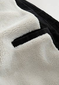 Close-up van beige fleece stof met een zwarte corduroy afwerking en een klein rechthoekig zwart zakdetail.