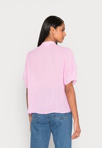 Camisa rosa de mangas curtas com padrão de bolinhas, corte descontraído, colarinho mandarim, tecido suave e detalhe de pregas nas costas, combinada com jeans azuis.