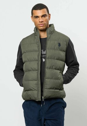 Homme portant un gilet matelassé vert olive sur une chemise noire à manches longues, les mains dans les poches du gilet, debout devant un fond clair uni.