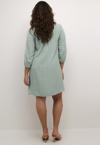 Hellgrünes Kleid mit drei Viertelärmeln, das ein dezentes vertikales Streifenmuster aufweist und einen lockeren Schnitt hat, der oberhalb des Knies endet.