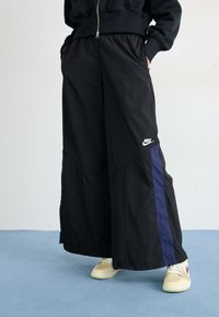 Svarta vida byxor med lila sidostrie, elastisk midja och Nike-logga, tillverkade av lätt, strukturerad textil.