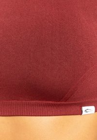 Close-up van een persoon die een geribbeld, donkerrood crop top draagt met een klein wit label aan de onderrand.