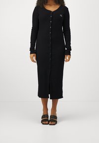 Pitkä musta mekko pehmeästä kankaasta, jossa on napitus edessä, pitkät hihat ja sivusääritys. Mekossa on myös pieni logo rinnassa.