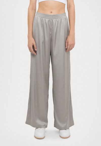 Lichtgrijze wijde broek van gladde stof met een elastische tailleband, kenmerkend door een losse pasvorm en een lichte glans. Gecombineerd met witte schoenen.