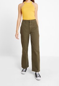 Top giallo a collo halter a coste abbinato a pantaloni a gamba larga verde oliva. Sneakers nere completano il look. L'outfit presenta linee e texture semplici.