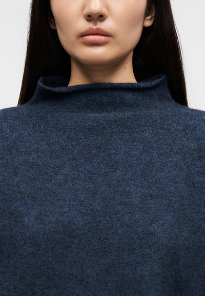Κοντινό πλάνο ενός ατόμου που φοράει σκούρο μπλε μάλλινο πουλόβερ με χαλαρή λαιμόκοψη, δείχνοντας μόνο το κάτω μέρος του προσώπου και τον λαιμό.