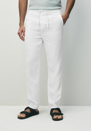 White linen pants with an elastic drawstring waist and side pockets, paired with black sandals featuring adjustable straps.