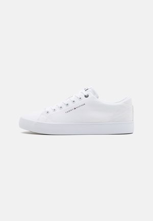 Hvid lavtop sneaker med hvide snørebånd, flad sål og lille Tommy Hilfiger-logo på siden mod en ensfarvet baggrund.