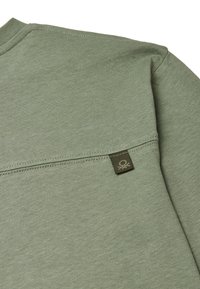 Zelená košile s dlouhým rukávem a texturovanou tkaninou, která má ševové detaily a malou hnědou nášivku na levé straně.