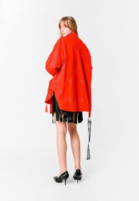 Camisa roja de gran tamaño con una textura suave y detalles bordados, combinada con una falda negra con flecos y zapatos de tacón alto negros.