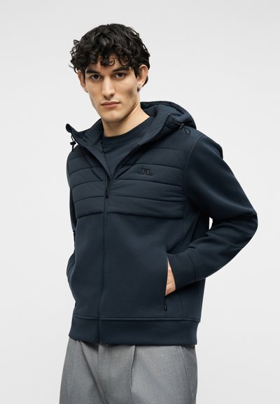 Άνδρας που φοράει μπλε σκούρο ζακέτα με φερμουάρ και κουκούλα, με επένδυση στον θώρακα, και γκρι παντελόνι, με τα χέρια στις τσέπες, μπροστά σε απλό φωτεινό φόντο.